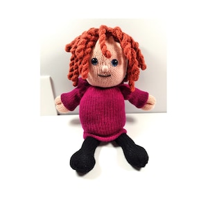 Op de afbeelding: Een gebreide pop met rood garen haar, een roze trui en een zwarte broek. De pop heeft blauwe ogen en een glimlachend gezicht.
