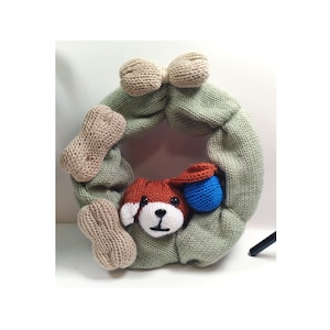 May include: A green knitted wreath with a brown dog face and two beige bone shapes. The wreath has a beige bow at the top and a blue ball.