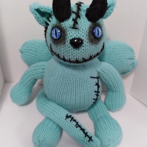 May include: A teal blue knitted plush toy with black horns, stitched seams, and blue eyes. The toy has a stitched mouth and a black nose.