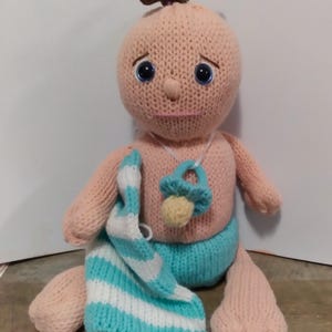 Peut inclure: Une poupée en tricot avec un corps rose clair, des yeux bleus et des cheveux bruns. La poupée porte une couverture rayée bleu et blanc et une couche en tricot bleue avec une sucette.