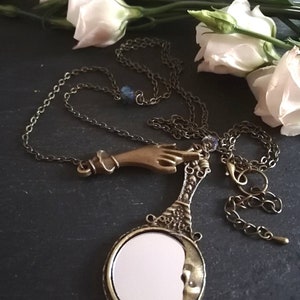 Puede incluir: Un collar de oro con un dije de mano y un dije de espejo en forma de media luna. El collar est&aacute; sobre un fondo oscuro con flores blancas.