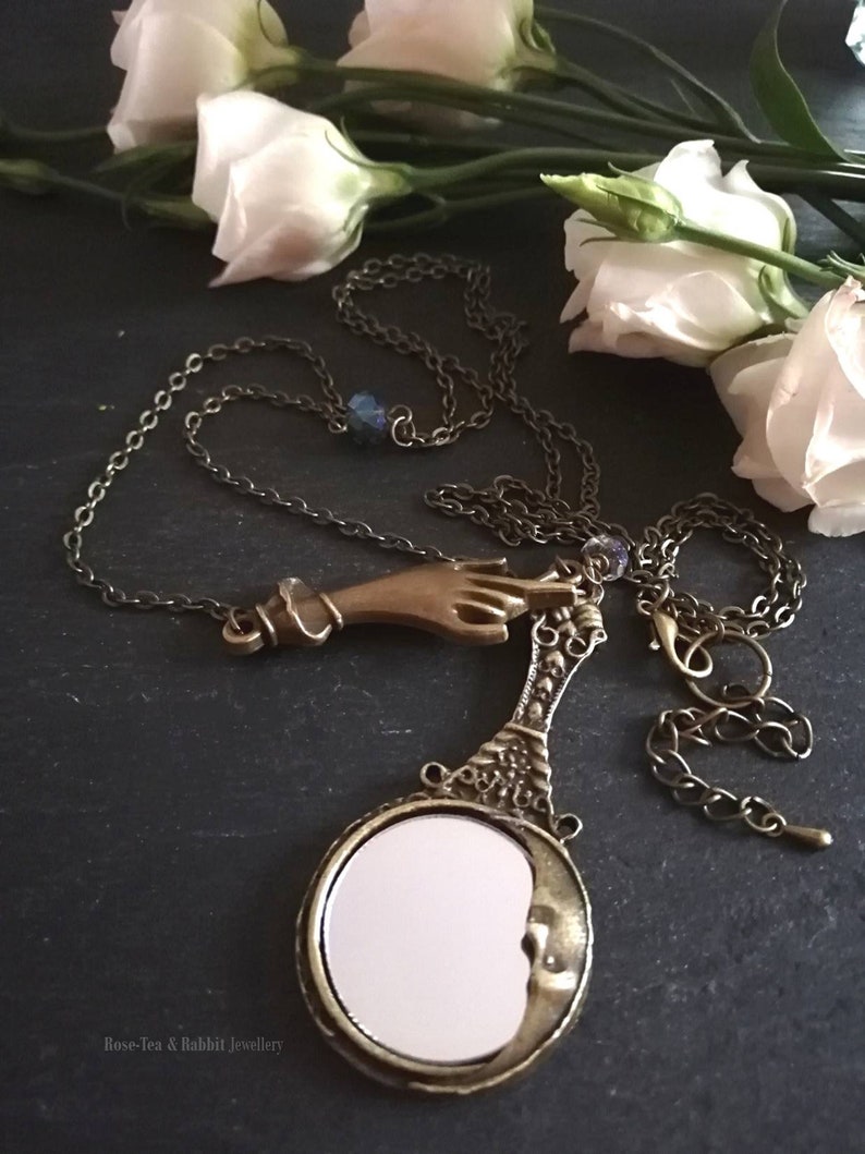Puede incluir: Un collar de oro con un dije de mano y un dije de espejo en forma de media luna. El collar est&aacute; sobre un fondo oscuro con flores blancas.