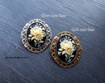 Puede incluir: Dos broches ovalados con un camafeo blanco con un diseño floral. Los broches tienen un fondo negro y un marco adornado en tono plateado o dorado. El texto "Silver-tone Base" y "Gold-tone Base" está por encima de los broches. El texto "Rose-Tea & Rabbit Jewellery" está debajo de los broches.