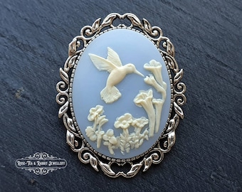 May include: Oval cameo brooch with a light blue background and a hummingbird design. The hummingbird is cream-colored, as are the flowers and foliage. The brooch has a silver-colored filigree frame. The Rose-Tea & Rabbit Jewellery logo is at the bottom.