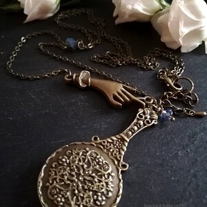 Puede incluir: Un collar de bronce con un dije de mano, un peque&ntilde;o espejo con un dise&ntilde;o intrincado y un dije de cristal azul. El collar est&aacute; sobre un fondo negro con flores blancas.