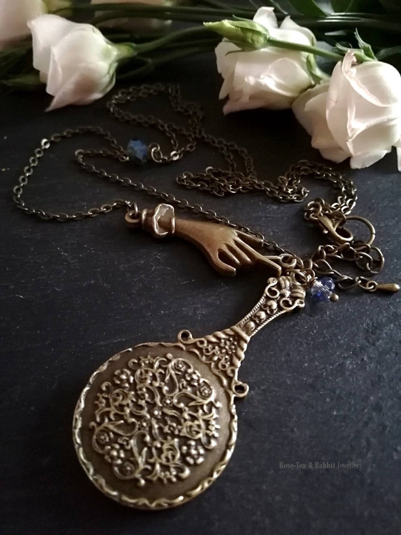 Puede incluir: Un collar de bronce con un dije de mano, un peque&ntilde;o espejo con un dise&ntilde;o intrincado y un dije de cristal azul. El collar est&aacute; sobre un fondo negro con flores blancas.