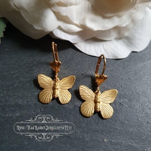 May include: A pair of gold-colored butterfly earrings. Each earring features a detailed butterfly pendant with textured wings and a small body. The earrings have a lever-back closure. The background includes a white flower and a dark gray surface.