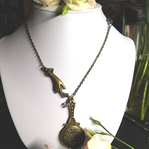Puede incluir: Un collar de oro con un dije de mano y un dije de espejo decorativo. El collar est&aacute; en un maniqu&iacute; blanco y hay flores blancas en el fondo.
