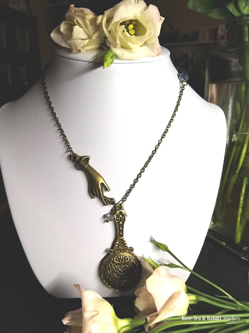 Puede incluir: Un collar de oro con un dije de mano y un dije de espejo decorativo. El collar est&aacute; en un maniqu&iacute; blanco y hay flores blancas en el fondo.