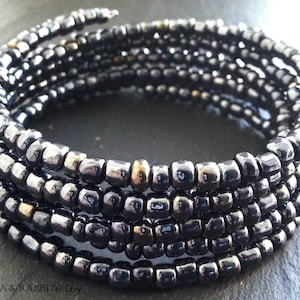 Könnte beinhalten: Ein Wickelarmband aus kleinen, dunkelgrauen Perlen. Das Armband wickelt sich um, wodurch mehrere Schichten entstehen. Einige goldfarbene Perlen sind eingestreut. Es ist ein Schmuckstück.