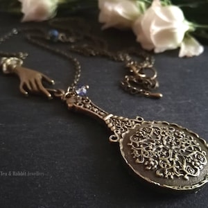 Puede incluir: Un collar dorado con un dije de mano y un colgante con forma de espejo de mano vintage con intrincados dise&ntilde;os florales. El espejo est&aacute; rodeado por un borde decorativo.