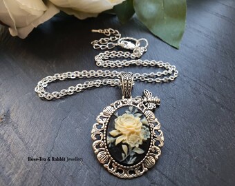 Puede incluir: Un collar de cadena de plata con un colgante de camafeo que presenta un diseño de flor tallada en blanco sobre un fondo negro. El colgante está rodeado por un marco de plata adornado y tiene un pequeño dije de mariposa de plata.