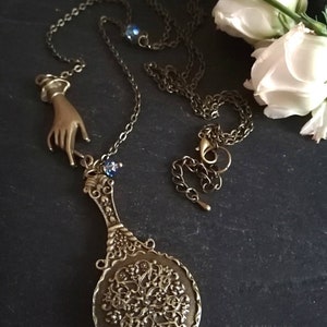 Puede incluir: Un collar de oro con un dije de mano y un dije de espejo decorativo. El espejo es redondo y tiene un dise&ntilde;o de filigrana. El collar est&aacute; sobre un fondo oscuro con flores blancas.