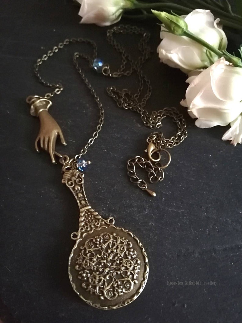 Puede incluir: Un collar de oro con un dije de mano y un dije de espejo decorativo. El espejo es redondo y tiene un dise&ntilde;o de filigrana. El collar est&aacute; sobre un fondo oscuro con flores blancas.