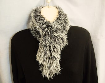 May include: A fluffy, textured scarf featuring a blend of black and white fibres, worn around the neck over a solid black long-sleeved shirt. The scarf has a shaggy, faux-fur appearance with a high-contrast colour pattern throughout the material.