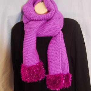 May include: A bright purple knitted scarf featuring a textured garter stitch pattern throughout the main body. The ends of the scarf are finished with a thick, shaggy trim in a vibrant, metallic magenta color that adds a distinct contrast.