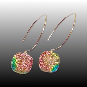 Pode incluir: Um par de brincos iridescentes com um pingente de vidro quadrado. Os brincos são feitos de metal dourado e têm um design simples e elegante.