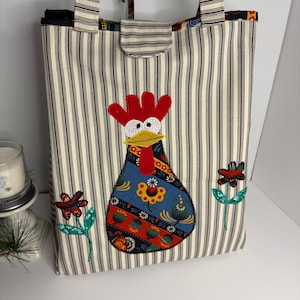 May include: A tote bag with a vertical black and white striped pattern. The bag features a colorful appliqué of a rooster and embroidered flowers. The handles and a small flap are also striped.
