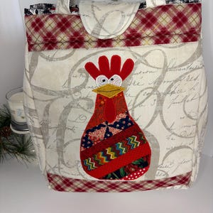 May include: A fabric tote bag with a rooster applique. The bag features a cream-colored background with script writing and red plaid accents. The rooster is made of various patterned fabrics in red, blue, and green.