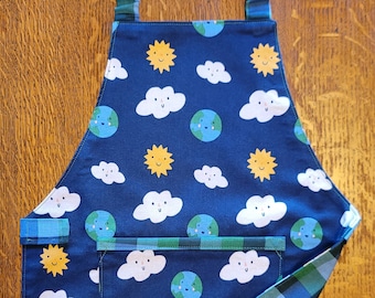 May include: A navy blue apron with a repeating pattern of smiling suns, clouds, and Earths. The apron has a pocket and a plaid pocket lining. The neck strap is elasticized with blue and green fabric.