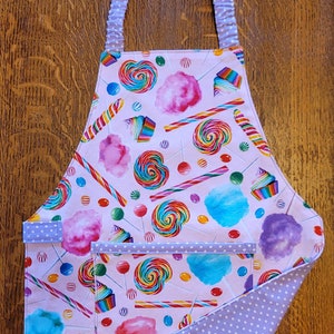 May include: A pink and purple candy-themed apron with a large pocket. The apron has a white polka dot trim and a white polka dot fabric on the back.