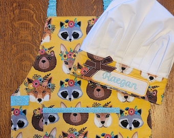 May include: A yellow children's apron with a blue trim and a white chef's hat. The apron has a pocket and is decorated with a repeating pattern of animal faces with floral crowns. The chef's hat has a brown bow and a personalized name tag that reads "Raegan".