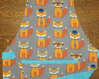May include: Gray apron with a repeating pattern of orange foxes wearing hats, glasses, and bow ties. The apron has a teal blue neck strap and pocket trim. The inside of the pocket and the back of the apron are teal blue.