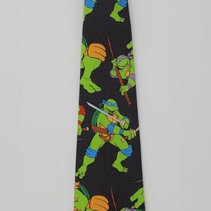 May include: A black and green necktie with a pattern of Teenage Mutant Ninja Turtles characters. The tie has a black adjustable strap.