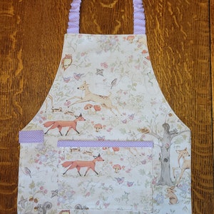 May include: A white apron with a woodland scene featuring deer, foxes, birds, and squirrels. The apron has a purple ruffle trim and a pocket with a purple ruffle trim.