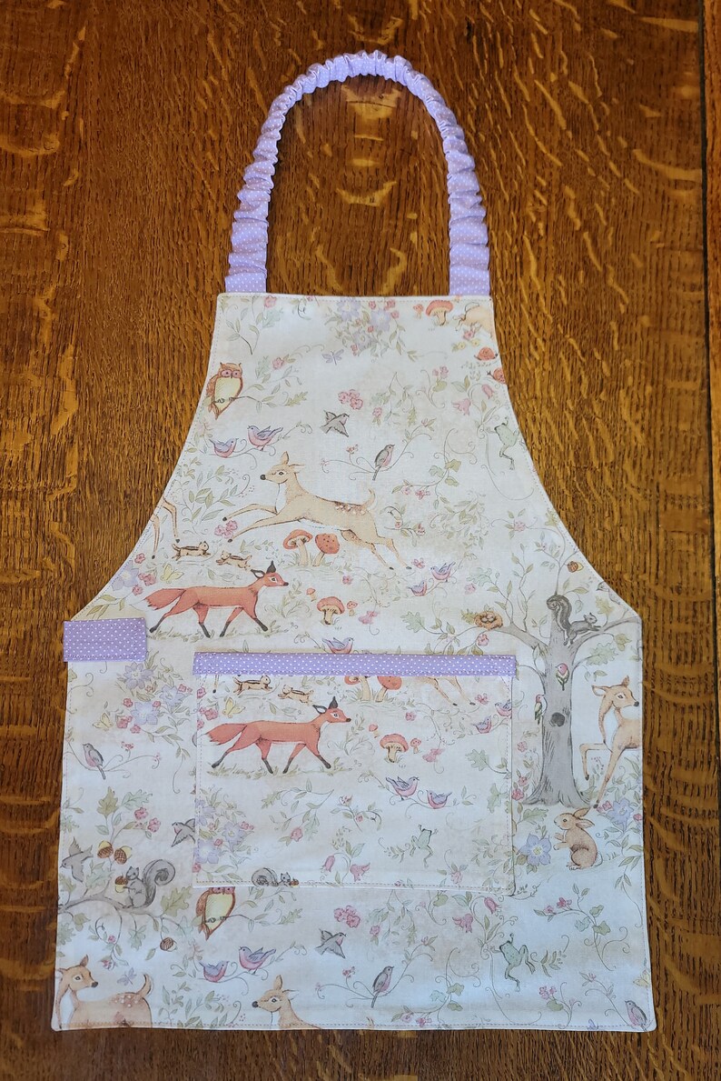 May include: A white apron with a woodland scene featuring deer, foxes, birds, and squirrels. The apron has a purple ruffle trim and a pocket with a purple ruffle trim.
