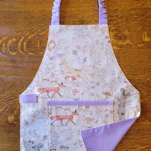May include: A white apron with a woodland print featuring deer, foxes, birds, and squirrels. The apron has a large pocket and purple polka dot trim.