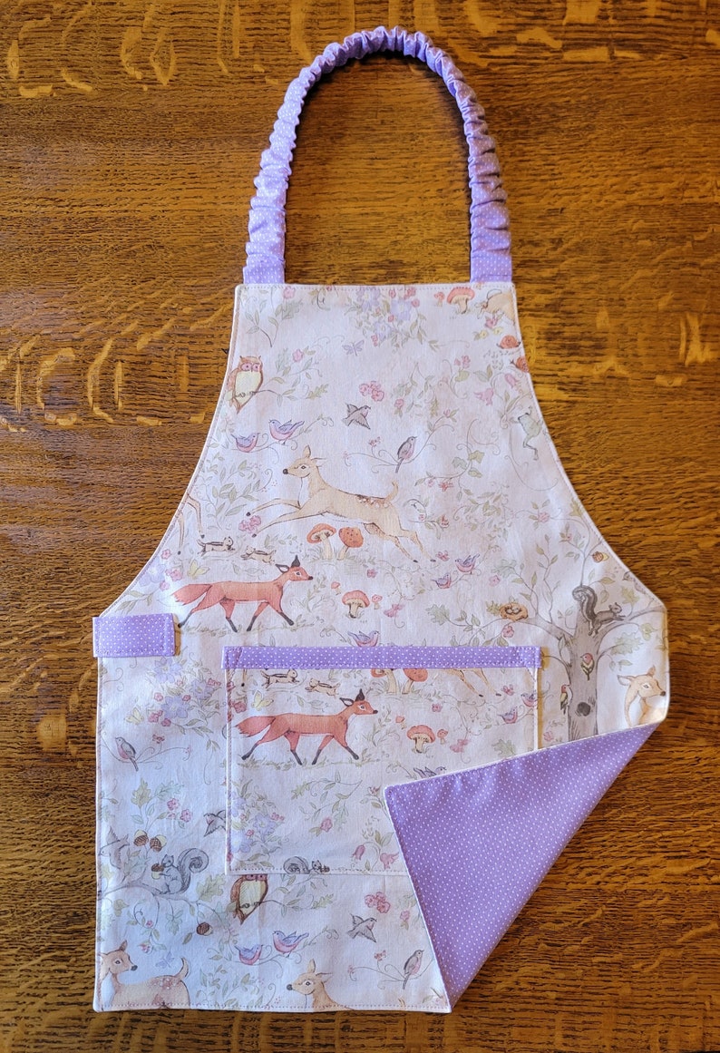 May include: A white apron with a woodland print featuring deer, foxes, birds, and squirrels. The apron has a large pocket and purple polka dot trim.