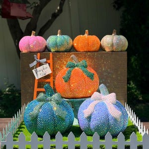 May include: A collection of six decorative pumpkins covered in sparkling rhinestones in various colors including pink, teal, orange, and iridescent white. The pumpkins feature fabric bows and are displayed with a small orange ladder and a sign reading Pumpkin Patch.