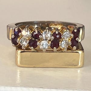 Può includere: Un anello in oro e argento con pietre preziose rosse e chiare alternate. L'anello è posto su una base rettangolare dorata. Il design dell'anello presenta una fascia di pietre preziose alternate.