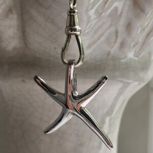 Può includere: Un ciondolo a forma di stella marina argentata è appeso a una delicata catena. La stella marina ha cinque punte e una superficie lucida e riflettente. La catena è attaccata a una chiusura, su sfondo bianco.