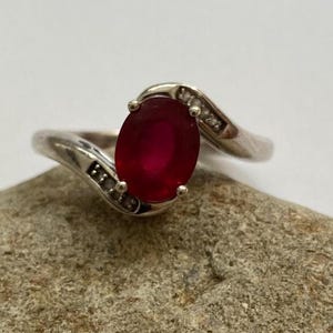 Op de afbeelding: Zilveren ring met een grote, ovale, dieprode edelsteen. De band heeft een sierlijke vorm, met kleine accentstenen. De ring wordt gepresenteerd tegen een neutrale achtergrond. De edelsteen is ongeveer 1,5 cm lang.