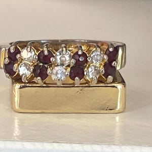 Puede incluir: Un anillo de oro con dos filas de piedras preciosas. La fila superior alterna entre piedras claras y rojo intenso. El anillo se asienta sobre una base rectangular dorada, creando un efecto en capas. Las piedras preciosas son redondas y están muy juntas.