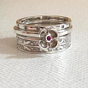 May include: A silver stackable ring set consisting of three bands. The top band features small circular indentations, the middle band displays a flower charm with a small red stone center, and the bottom band has an embossed leaf pattern.