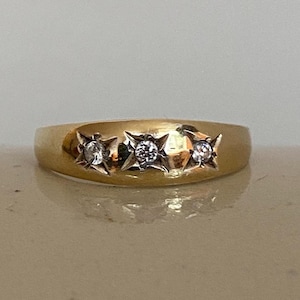 May include: A gold band ring featuring three small, clear, round-cut stones set in individual star-shaped metal engravings. The band has a smooth, polished finish and a rounded profile, showcasing a consistent yellow gold tone throughout the piece.