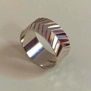Puede incluir: Un anillo de banda ancha de plata con un patrón repetitivo en forma de chevron o V grabado en la superficie exterior. El interior de la banda es liso y muestra claramente la palabra SILVER estampada en el metal, indicando la composición del material.