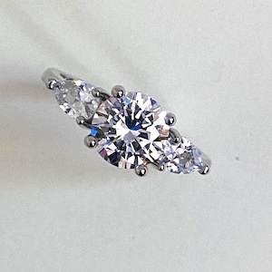 May include: A silver-toned ring featuring a large, round, brilliant-cut clear stone at the center, flanked by two smaller, pear-shaped clear stones on either side. The stones are held in place by metal prongs on a polished, reflective metal band.