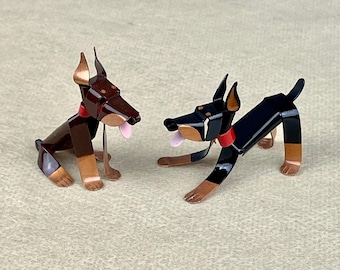 May include: Two metal Doberman Pinscher dog figurines. One is brown and black, the other is black. Both have red collars and are posed in a playful stance.