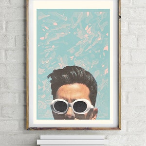May include: A framed art print featuring a stylized portrait of a person wearing white sunglasses. The background is a light blue with abstract pink and white shapes. The frame is a light brown color.
