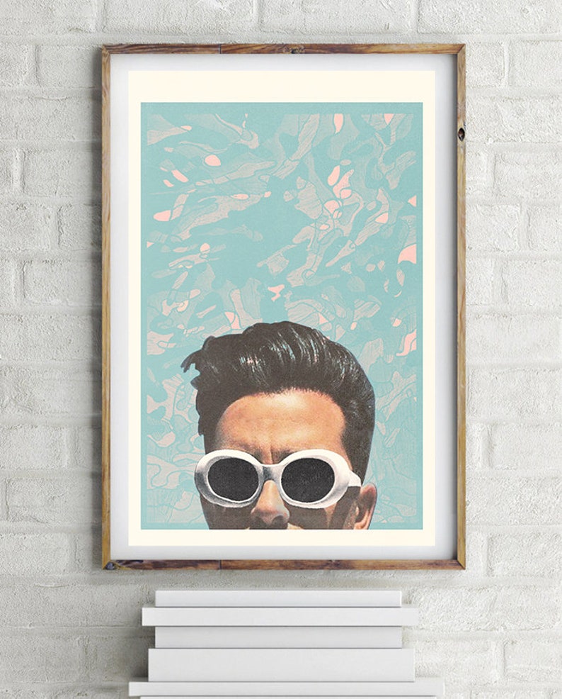 May include: A framed art print featuring a stylized portrait of a person wearing white sunglasses. The background is a light blue with abstract pink and white shapes. The frame is a light brown color.