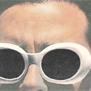 May include: Close-up of a person wearing white-framed sunglasses with large, oval black lenses. The image has a vintage, slightly faded aesthetic, with a light blue floral pattern in the background.