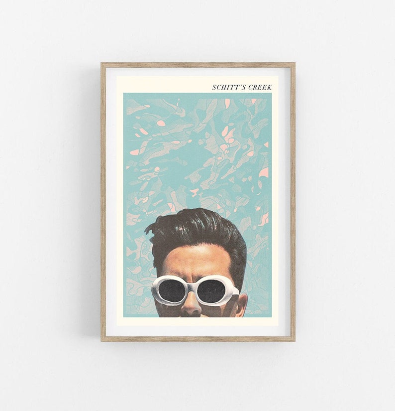 May include: A framed art print featuring a stylized image of a person wearing white sunglasses. The print has a light blue and pink abstract background. The words "SCHITT'S CREEK" are printed at the top of the print.