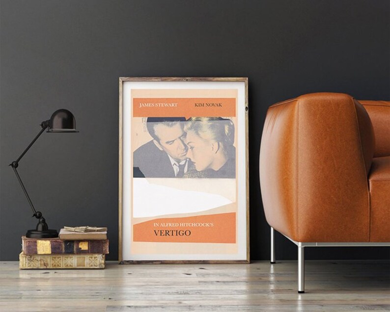 Puede incluir: Un p&oacute;ster enmarcado de la pel&iacute;cula "Vertigo" de Alfred Hitchcock con James Stewart y Kim Novak. El p&oacute;ster tiene un dise&ntilde;o vintage con acentos naranjas y blancos. Una l&aacute;mpara de escritorio negra y una pila de libros est&aacute;n a la izquierda, y un sof&aacute; de cuero marr&oacute;n est&aacute; a la derecha.