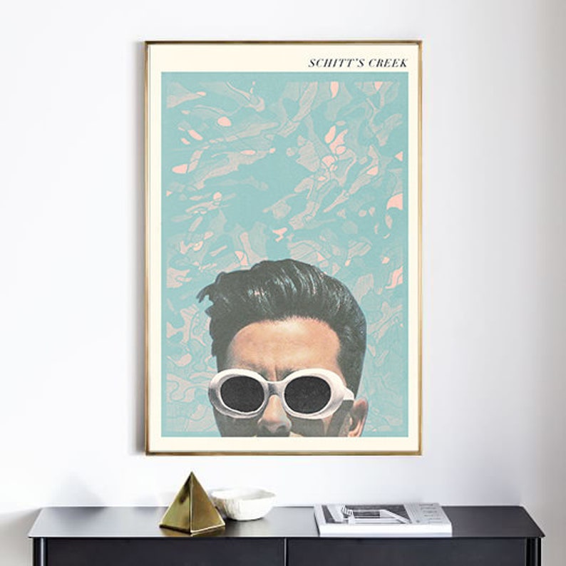 May include: A framed art print featuring a stylized image of a person wearing white sunglasses, with the text "SCHITT'S CREEK" at the top. The background is a mix of blue and pink hues. The print is in a gold frame.