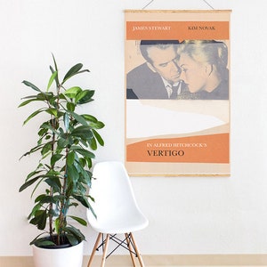 Puede incluir: Un p&oacute;ster enmarcado de la pel&iacute;cula "Vertigo" de Alfred Hitchcock con James Stewart y Kim Novak. El p&oacute;ster tiene un dise&ntilde;o vintage con tonos naranja y beige. Una silla blanca y una planta en maceta est&aacute;n en primer plano.