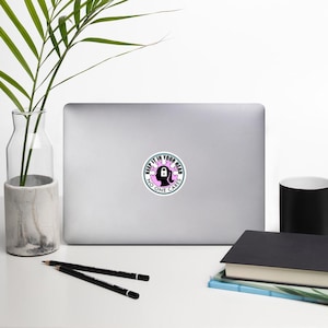 May include: A white laptop with a sticker on it. The sticker is a circle with a pink background and a black silhouette of a head with a lock on it. The text on the sticker reads "Keep it in your head no one cares".